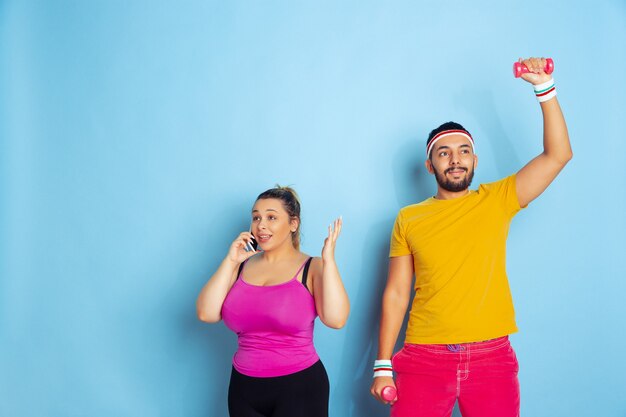 Jovem casal muito caucasiano com roupas brilhantes, treinando sobre fundo azul Conceito de esporte, emoções humanas, expressão, estilo de vida saudável, relação, família. Ele está treinando, ela está falando no telefone.