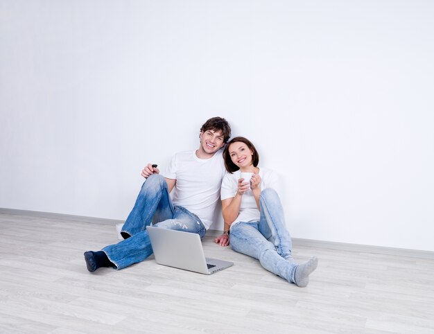 Jovem casal relaxando sentado em uma sala vazia com laptop e bebendo chá