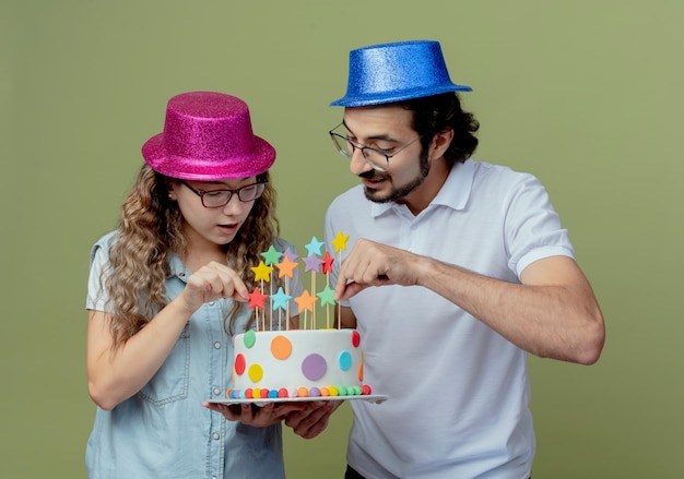 Jovem casal satisfeito com um chapéu rosa e azul, segurando e olhando para um bolo de aniversário