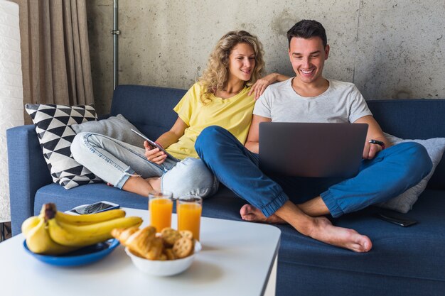 Jovem casal sentado no sofá em casa, usando dispositivos digitais, segurando um laptop