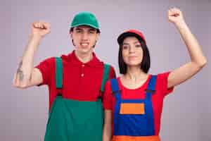 Foto grátis jovem casal sorridente cara feliz garota com uniforme de trabalhador da construção civil e boné olhando para a câmera fazendo gesto de bater isolado na parede branca