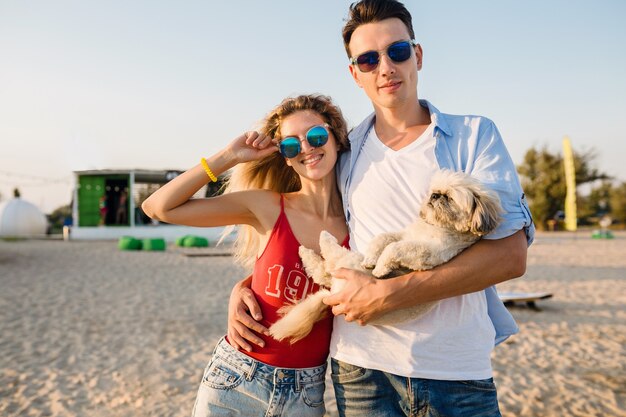 Jovem casal sorridente e atraente se divertindo na praia brincando com o cachorro da raça shih-tsu