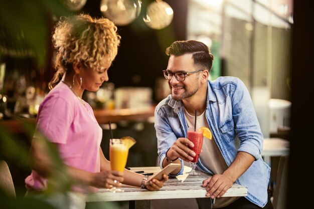 Jovem casal sorridente relaxando e bebendo coquetéis de frutas em um café Mulher está enviando mensagens de texto no celular