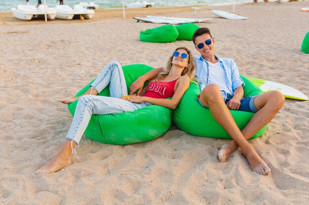 Jovem casal sorridente se divertindo na praia, sentado na areia com pranchas de surf