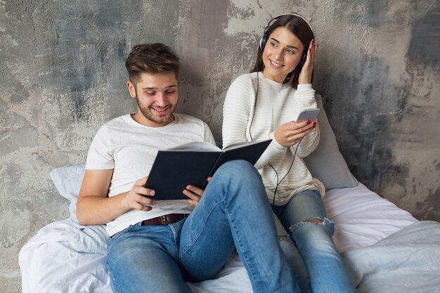 Jovem casal sorridente, sentado na cama em casa com roupa casual, lendo um livro de jeans, um homem lendo um livro, uma mulher ouvindo música em fones de ouvido e passando um tempo romântico juntos