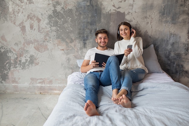 Jovem casal sorridente, sentado na cama em casa com roupa casual, lendo um livro de jeans, um homem lendo um livro, uma mulher ouvindo música em fones de ouvido e passando um tempo romântico juntos