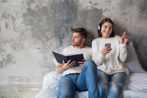 Jovem casal sorridente, sentado na cama em casa com roupa casual, lendo um livro de jeans, um homem lendo um livro, uma mulher ouvindo música em fones de ouvido e passando um tempo romântico juntos