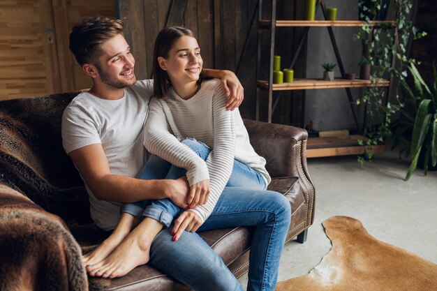 Jovem casal sorridente, sentado no sofá em casa com roupa casual, amor e romance, mulher e homem se abraçando, vestindo jeans, relaxando juntos