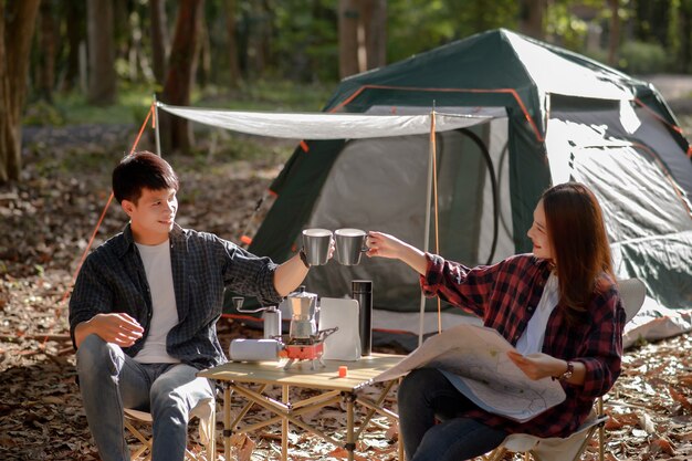 Jovem casal tilintando canecas de café pela manhã em frente a uma barraca de acampamento pela manhã no parque natural