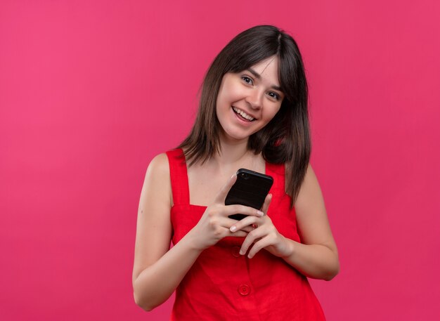 Jovem caucasiana alegre segurando o telefone com as duas mãos e olhando para a câmera em um fundo rosa isolado com espaço de cópia