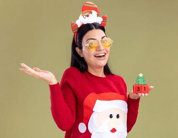 Jovem caucasiana alegre usando bandana de Papai Noel e suéter com óculos segurando um brinquedo de árvore de Natal com data olhando para a câmera.