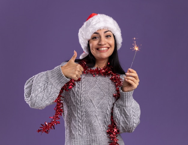 Jovem caucasiana alegre usando chapéu de Natal e guirlanda de ouropel no pescoço segurando um diamante de feriado aparecendo o polegar isolado na parede roxa