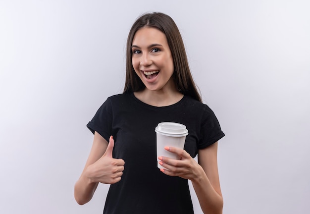 Jovem caucasiana alegre vestindo uma camiseta preta segurando uma xícara de café com o polegar para cima na parede branca isolada