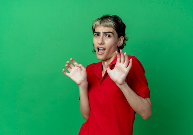 Jovem caucasiana assustada com corte de cabelo de duende, mostrando as mãos vazias, isoladas em um fundo verde com espaço de cópia