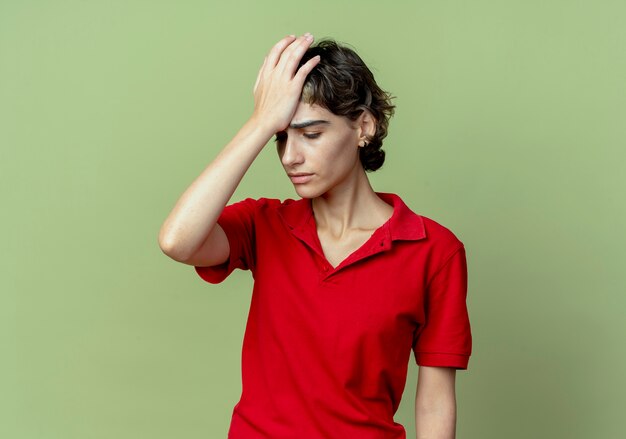 Jovem caucasiana cansada com corte de cabelo de duende, colocando a mão na cabeça, olhando para baixo, isolada em um fundo verde oliva com espaço de cópia