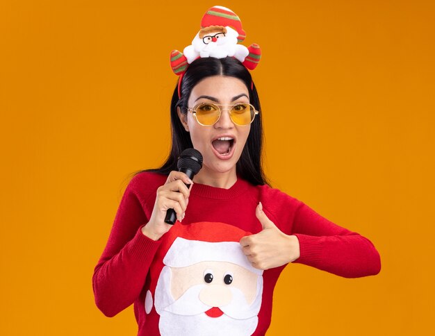 Jovem caucasiana confiante usando bandana de Papai Noel e suéter com óculos falando no microfone, olhando para a câmera aparecendo polegar isolado em fundo laranja