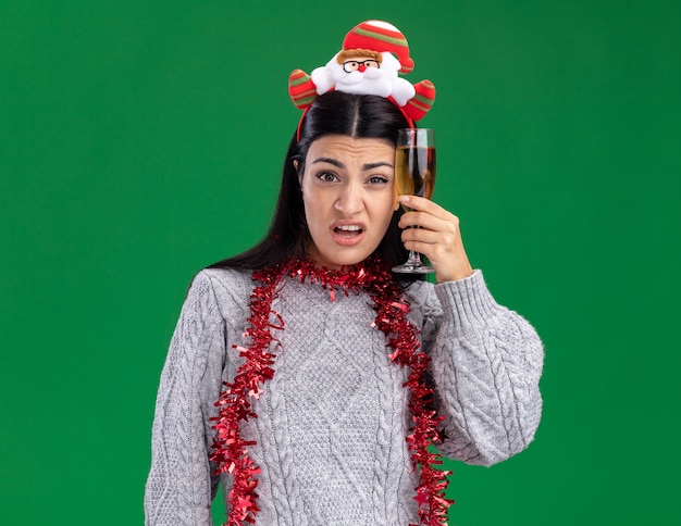 Foto grátis jovem caucasiana confusa usando bandana de papai noel e guirlanda de ouropel no pescoço segurando uma taça de champanhe, tocando a cabeça com ela isolada na parede verde