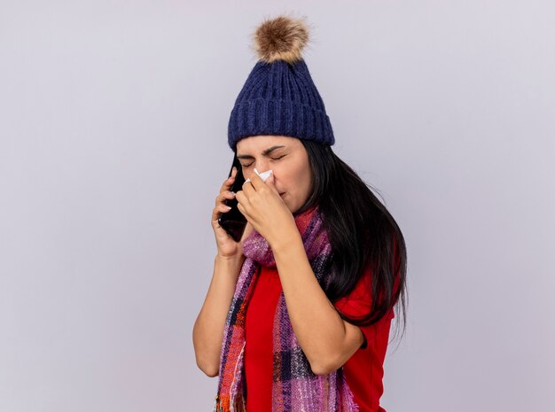 Jovem caucasiana doente, carrancuda, usando chapéu de inverno e lenço em pé em vista de perfil, falando no telefone, limpando o nariz com guardanapo