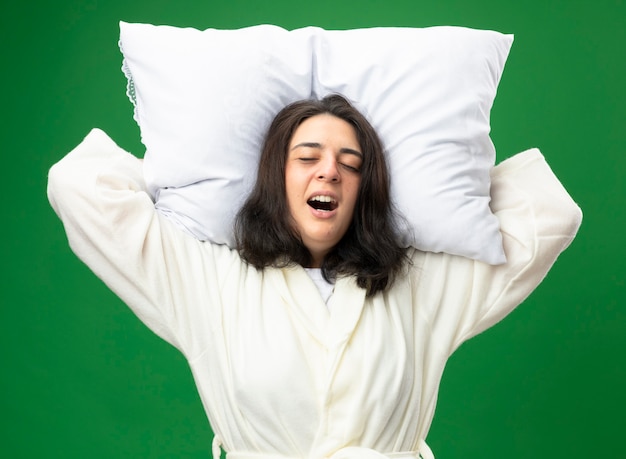 Foto grátis jovem caucasiana doente, dolorida, vestindo um manto, segurando o travesseiro sob a cabeça, gritando com os olhos fechados, isolado no fundo verde