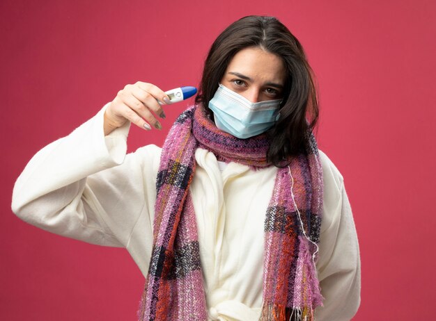 Jovem caucasiana doente e confiante usando manto e lenço com máscara segurando um termômetro isolado na parede vermelha