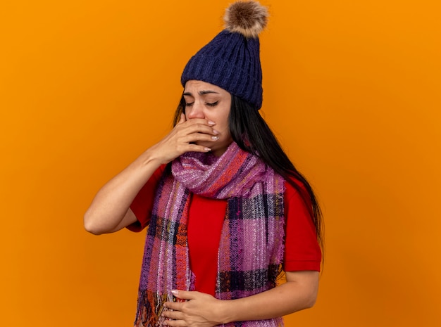 Foto grátis jovem caucasiana doente e dolorida, usando chapéu e lenço de inverno, mantendo a mão na barriga e na boca, preparando-se para espirrar com os olhos fechados, isolada na parede laranja com espaço de cópia