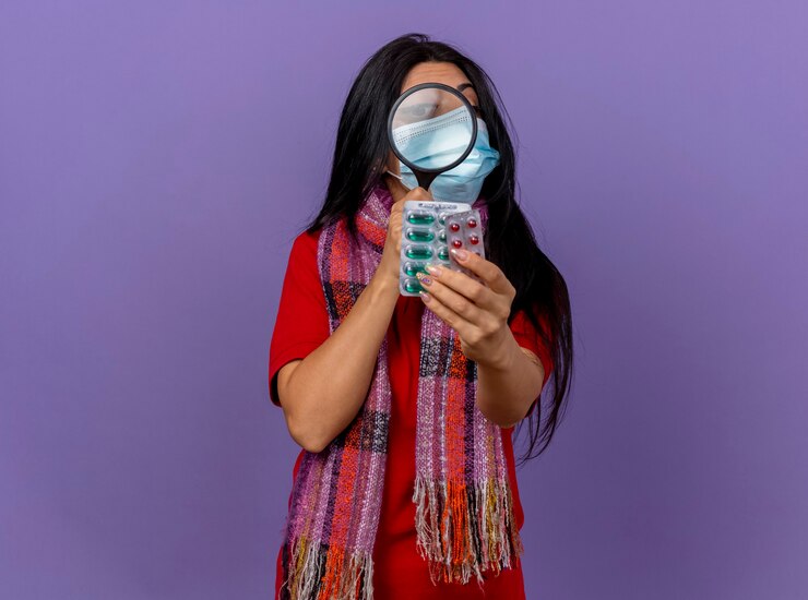 Foto grátis jovem caucasiana doente usando máscara e lenço estendendo pacotes de cápsulas, olhando para eles através de uma lupa isolada na parede roxa com espaço de cópia
