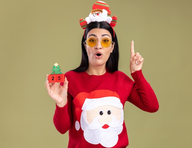 Jovem caucasiana impressionada com bandana de Papai Noel e suéter com óculos segurando um brinquedo de árvore de Natal com data apontando para cima isolada na parede verde oliva