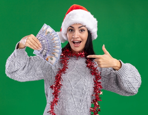 Foto grátis jovem caucasiana impressionada com chapéu de natal e guirlanda de ouropel no pescoço segurando e apontando para o dinheiro isolado na parede verde