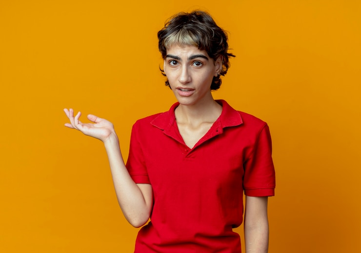 Foto grátis jovem caucasiana impressionada com corte de cabelo de duende mostrando a mão vazia isolada em um fundo laranja com espaço de cópia