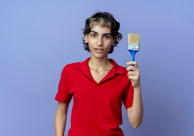 Foto grátis jovem caucasiana impressionada com corte de cabelo de duende segurando um pincel isolado em um fundo roxo com espaço de cópia