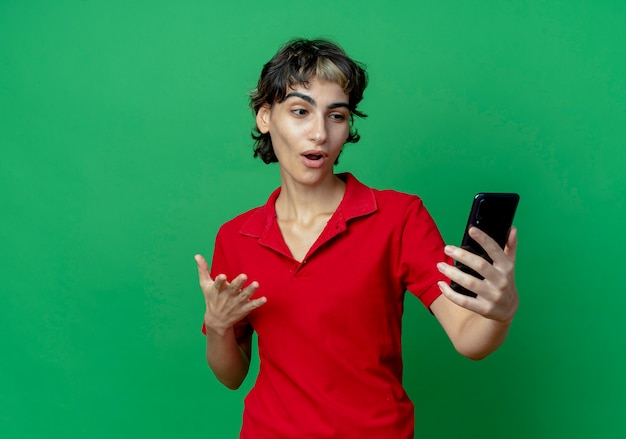 Jovem caucasiana impressionada com um corte de cabelo de duende segurando e olhando para o telefone celular e mantendo a mão no ar