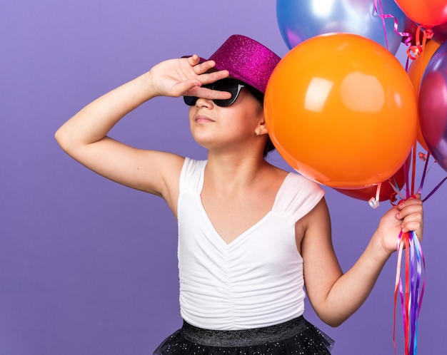 Jovem caucasiana insatisfeita com óculos de sol com chapéu de festa violeta segurando balões de hélio e mantendo a mão na frente do rosto isolada na parede roxa com espaço de cópia