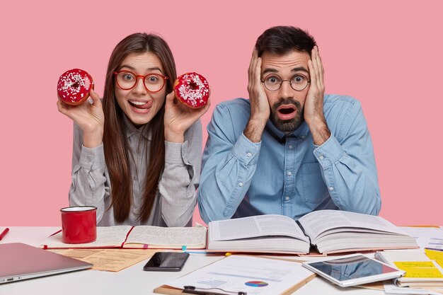 jovem caucasiana positiva come rosquinhas vermelhas saborosas, usa óculos, frustrada e intrigada cara com a barba por fazer em uma camisa formal se sente farta do trabalho