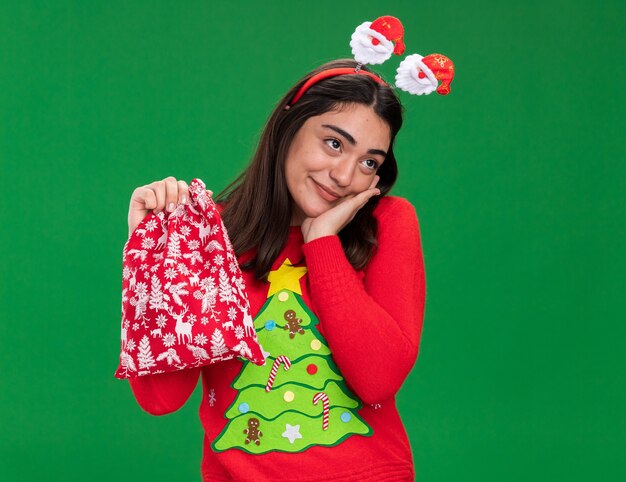 Jovem caucasiana satisfeita com fita de Papai Noel coloca a mão no rosto e segura uma sacola de presente de Natal isolada em um fundo verde com espaço de cópia