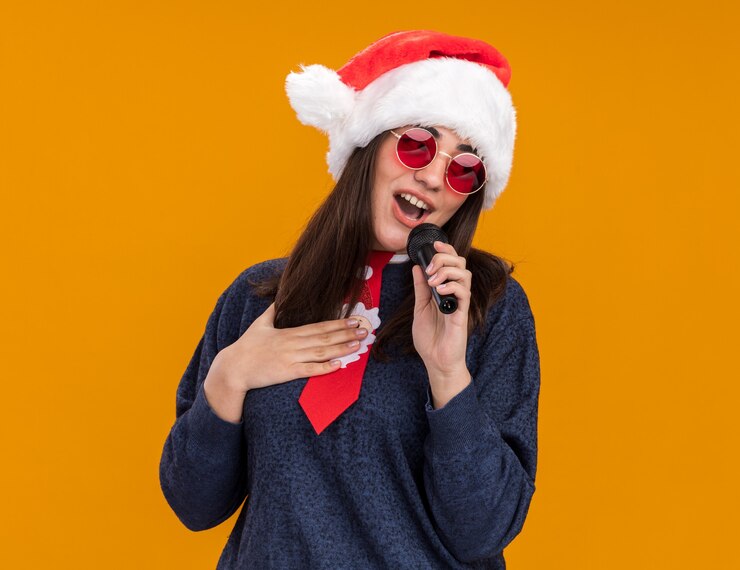 Foto grátis jovem caucasiana satisfeita com óculos de sol com chapéu de papai noel e gravata de papai noel coloca a mão no peito e segura o microfone fingindo cantar
