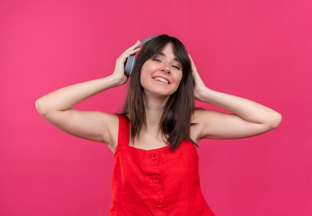 Jovem caucasiana satisfeita segurando fones de ouvido na cabeça em fundo rosa isolado