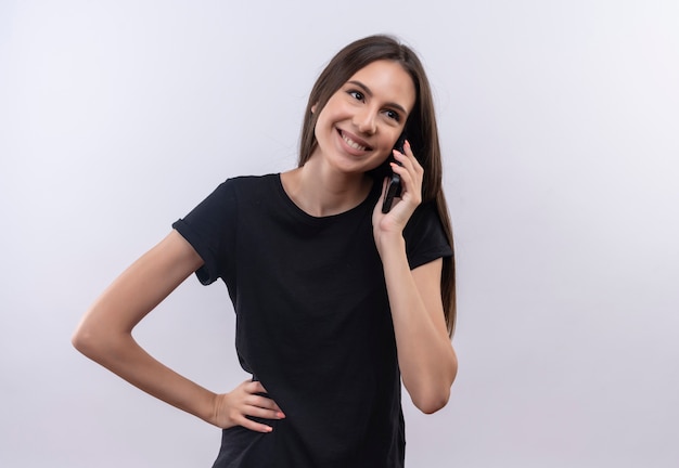 Jovem caucasiana sorridente, vestindo uma camiseta preta, falando ao telefone, colocando a mão no quadril no fundo branco isolado