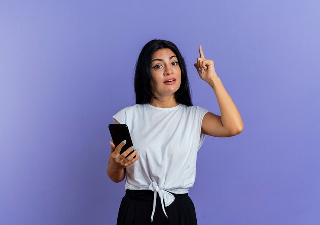 Jovem caucasiana surpreendida segurando o telefone e apontando para cima