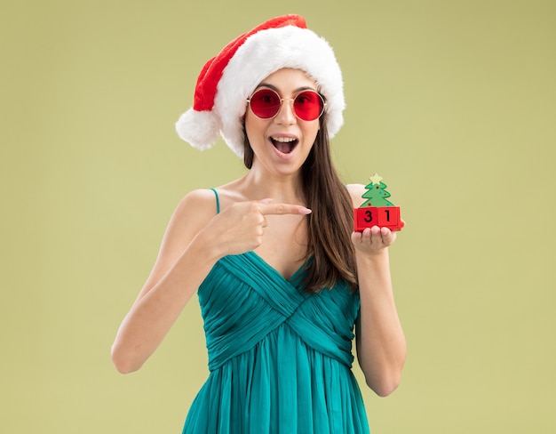 Jovem caucasiana surpresa com óculos de sol e chapéu de Papai Noel segurando e apontando para um enfeite de árvore de Natal