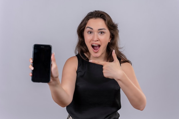 Foto grátis jovem caucasiana surpresa, vestindo uma camiseta preta, estendendo o telefone para a frente e apontando para o telefone no fundo branco