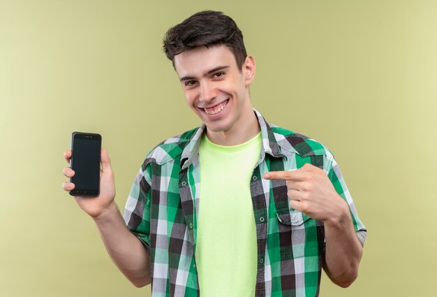 Jovem caucasiano sorridente com camisa verde aponta para o telefone na mão em fundo verde isolado