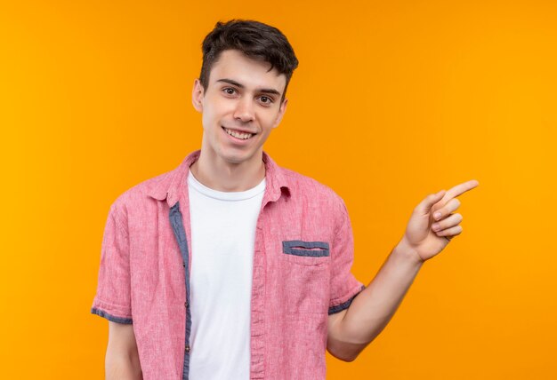 Jovem caucasiano sorridente, vestindo uma camisa rosa, aponta para o lado em um fundo laranja isolado