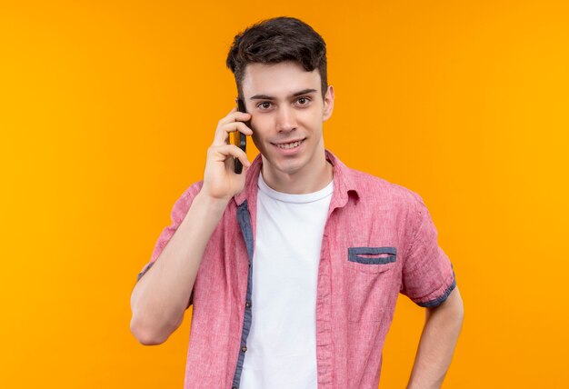 Jovem caucasiano sorridente, vestindo uma camisa rosa, fala ao telefone em um fundo laranja isolado