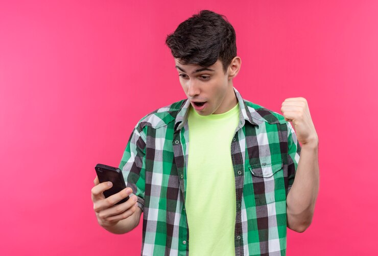 Foto grátis jovem caucasiano surpreso com uma camisa verde, segurando e olhando no telefone, mostrando um gesto de sim no fundo rosa isolado