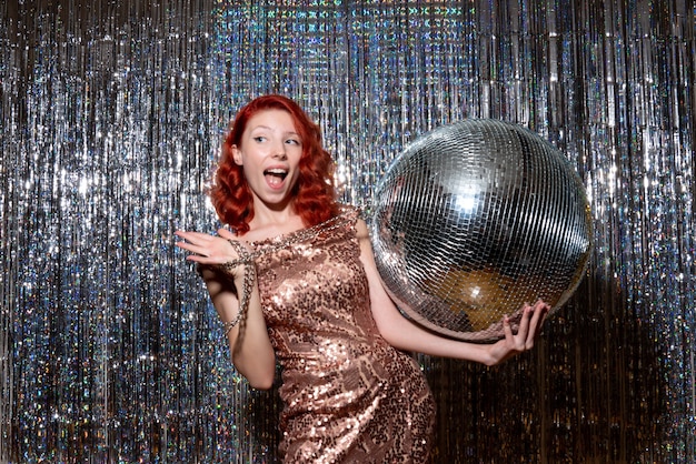 Jovem celebrando o ano novo em festa segurando uma bola de discoteca nas cortinas brilhantes