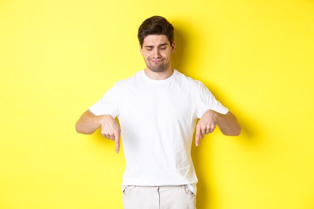 Jovem cético em camiseta branca, apontando e olhando para baixo, chateado, desaprova e não gosta do produto, de pé sobre um fundo amarelo.