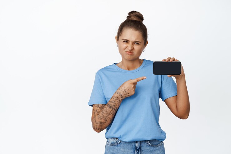 Foto grátis jovem chateada apontando o dedo para a tela do celular, smartphone horizontal, franzindo a testa chateada com o aplicativo, fundo branco