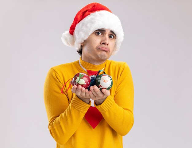 Jovem chateado com gola alta amarela e Papai Noel segurando bolas de natal, olhando para a câmera, franzindo os lábios em pé sobre um fundo branco