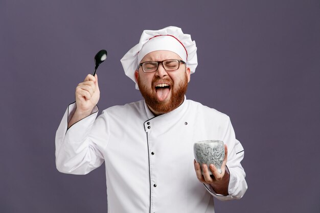 Jovem chef enojado vestindo uniforme de óculos e boné segurando a tigela mostrando a colher e a língua com os olhos fechados, isolados no fundo roxo