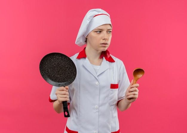 Jovem chef feminina loira ansiosa com uniforme de chef segurando uma frigideira e uma colher de pau, olhando para o lado isolado na parede rosa